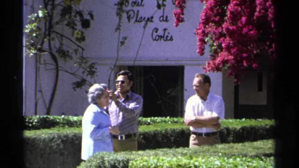 The 1975 Guaymas home-movie frame shows the flower placement nearing completion beside clipped hedges and a white Playa de Cortes wall.