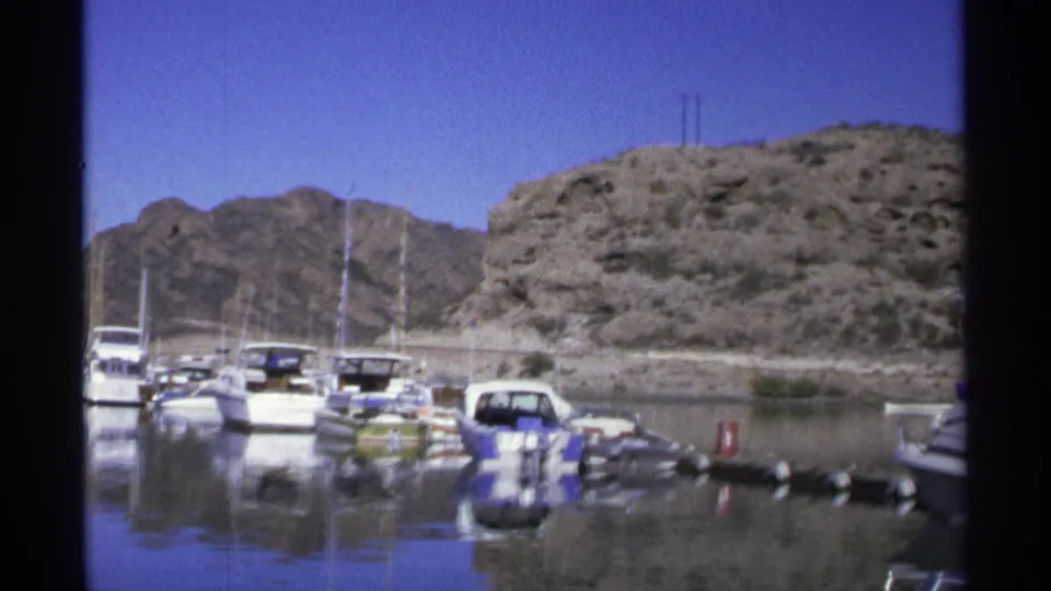1975 Guaymas marina view with small boats, sailboat masts, calm water, and a rocky hillside under blue sky.