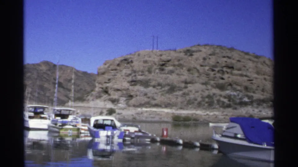 1975 Guaymas marina frame showing moored boats, a blue canvas cover, calm water, and rocky hillside terrain.