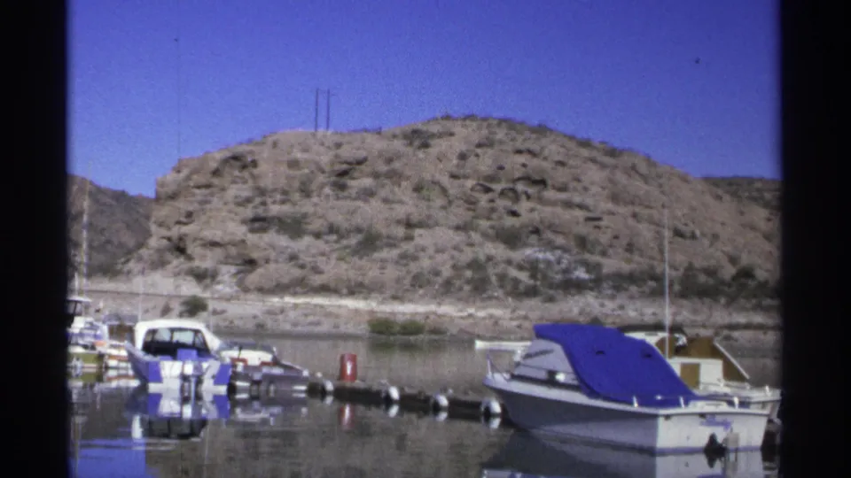 1975 Guaymas home-movie view of docked small boats, floating markers, still water, and sunlit rocky hillside.