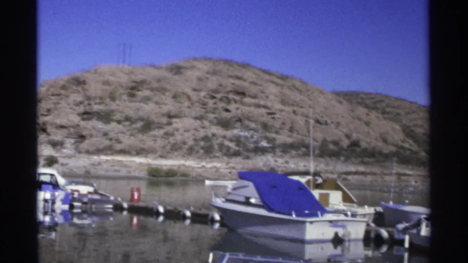 1975 Guaymas marina scene with a blue-covered motorboat in the foreground and rocky shoreline behind it.
