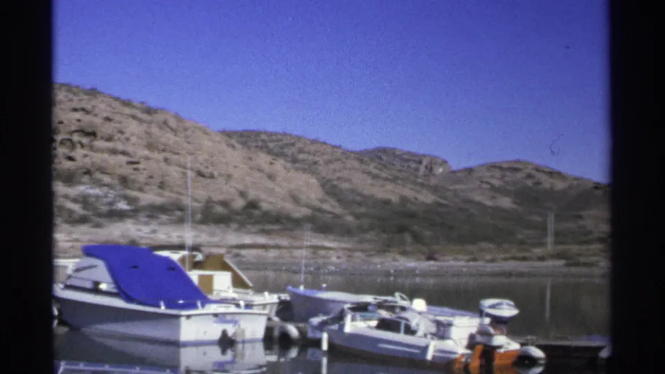 1975 home-movie frame from Guaymas with docked small boats, a cropped blue-covered boat, arid hills, and a quiet shoreline.