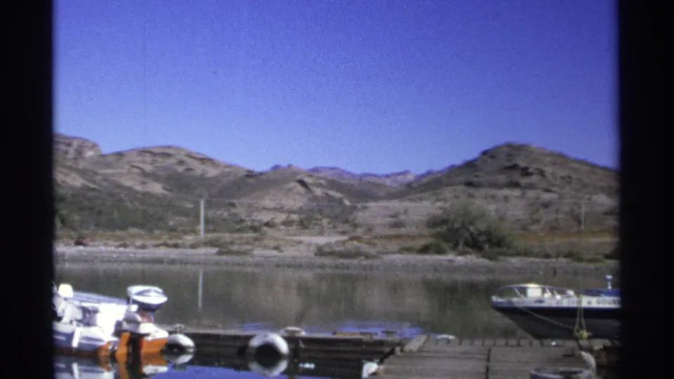 1975 8mm view of a Guaymas dock with small motorboats in the foreground, calm marina water, sparse brush, and rocky hills beyond.