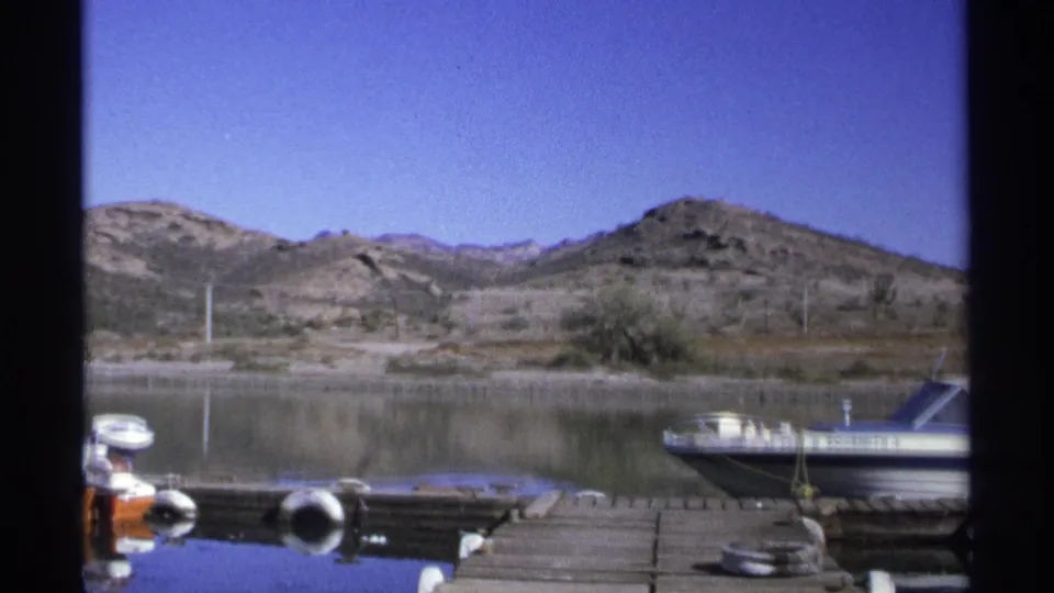 Guaymas marina frame from 1975 with a larger moored boat at right, dock hardware in front, reflective water, and arid hills beyond.