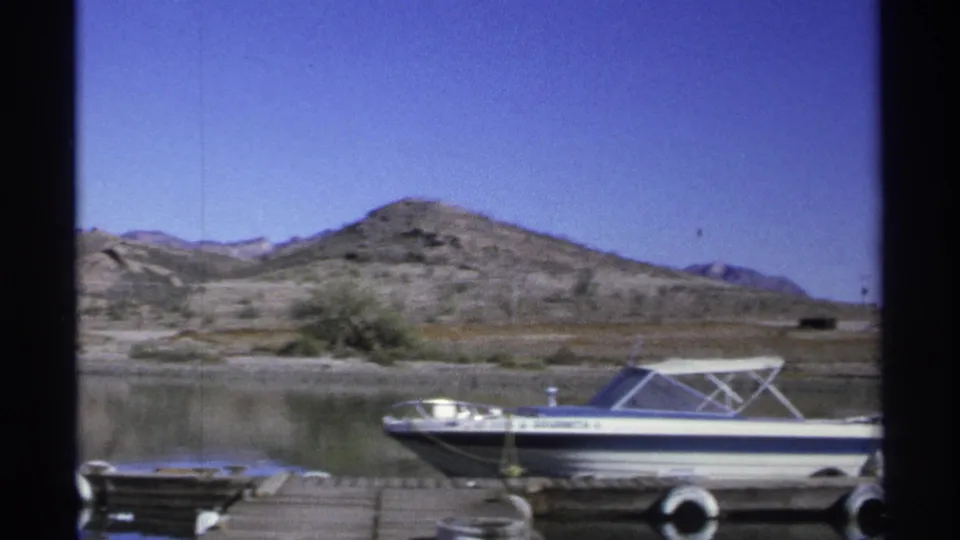 1975 Guaymas frame with the docked motorboat seen closer, its side lettering visible against calm water and dry hillside terrain.