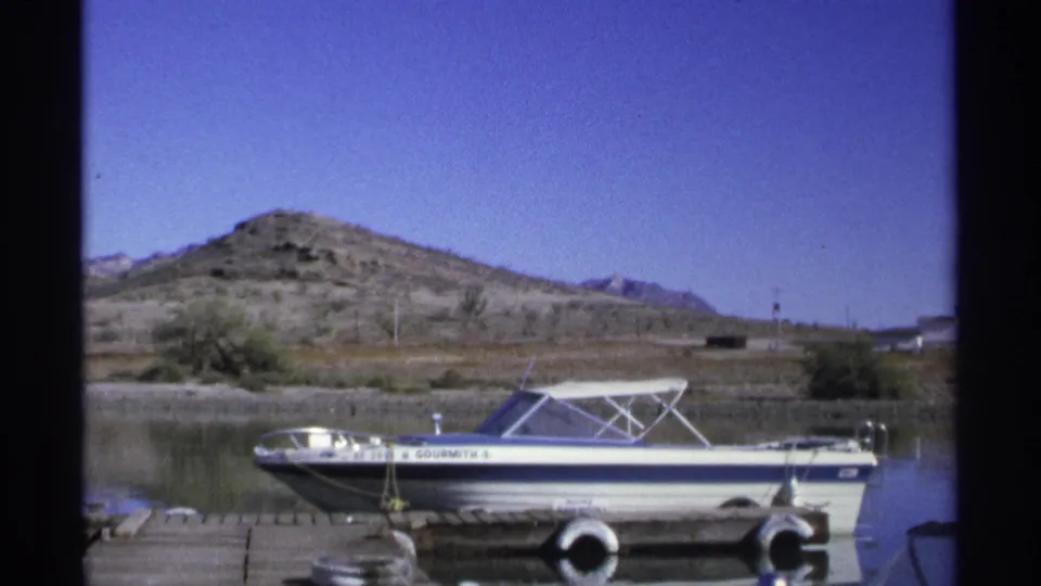 A 1975 Guaymas home-movie view of the motorboat broadside at the pier, with still water, shoreline brush, and rocky hills in the background.