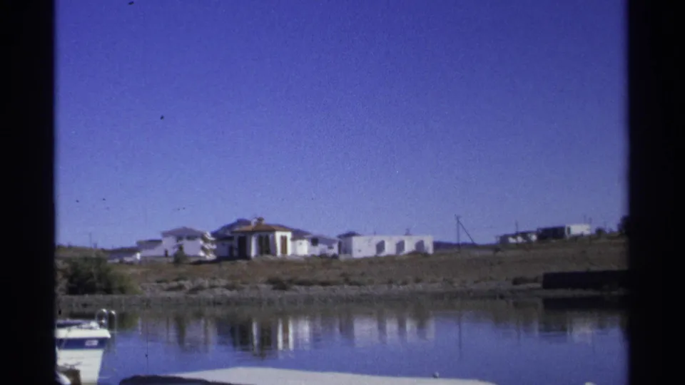 The final 1975 Guaymas frame opens to a wider shoreline view with calm water, partial boat, white buildings, utility poles, and open sky.