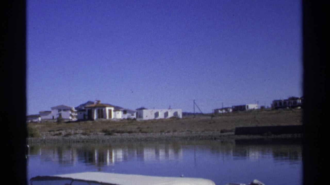 White shoreline buildings reflected in calm water under a blue sky.