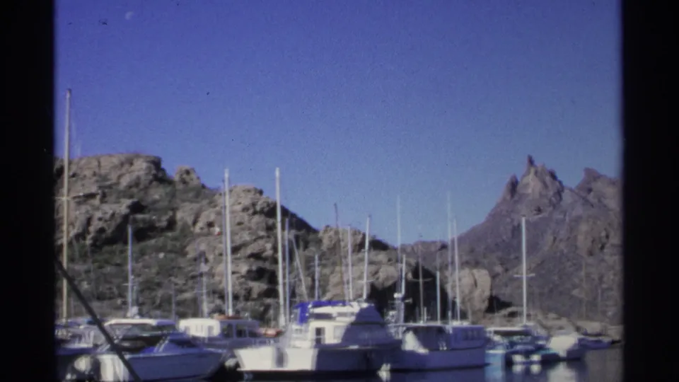 1975 Guaymas harbor view with docked boats, mast lines, rocky hills, and clear blue sky in 8mm color.