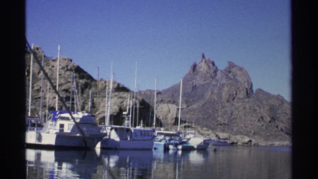 Docked boats and sailboat masts in front of rocky hills.