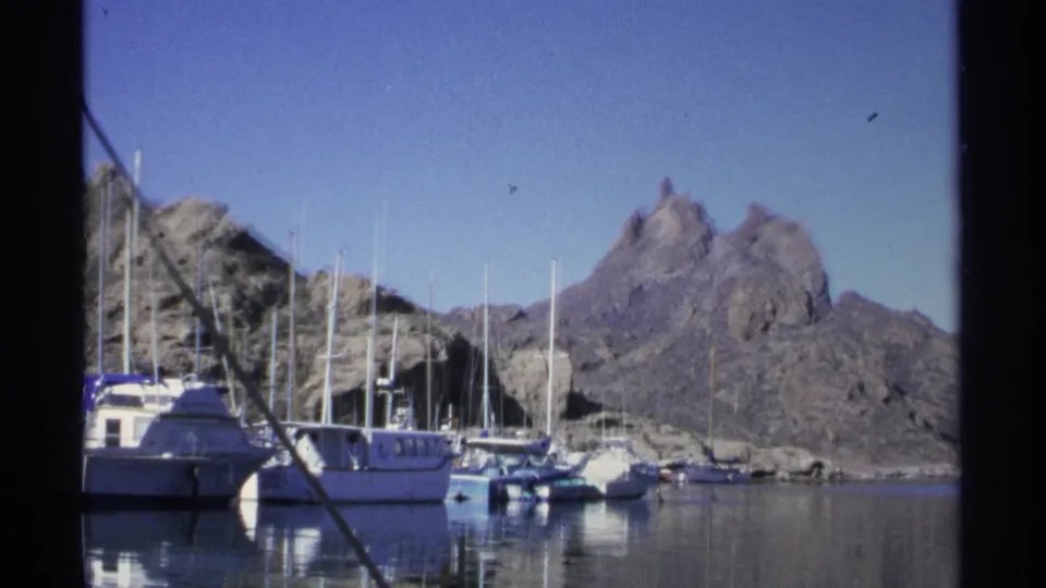 Vintage 8mm frame of a Guaymas marina with moored boats, masts, reflections, rocky shoreline, and dry mountains beyond.