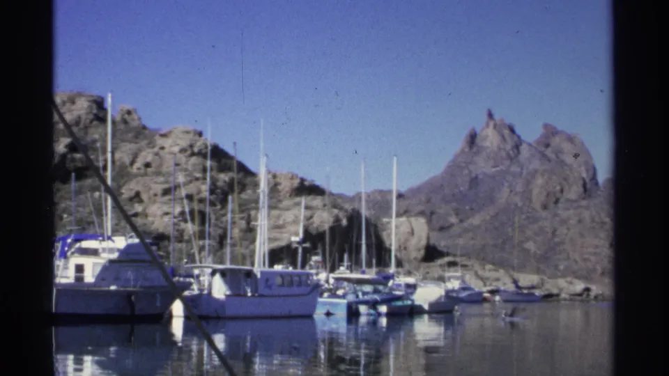 1975 home-movie frame from Guaymas showing docked boats, sail masts, still marina water, and steep rocky mountains under blue sky.