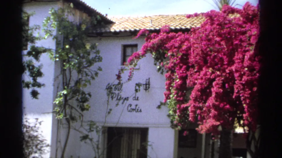 1975 home-movie frame of the Hotel Playa de Cortes exterior, with sign, tiled roof, shaded window, vines, and dense bougainvillea.