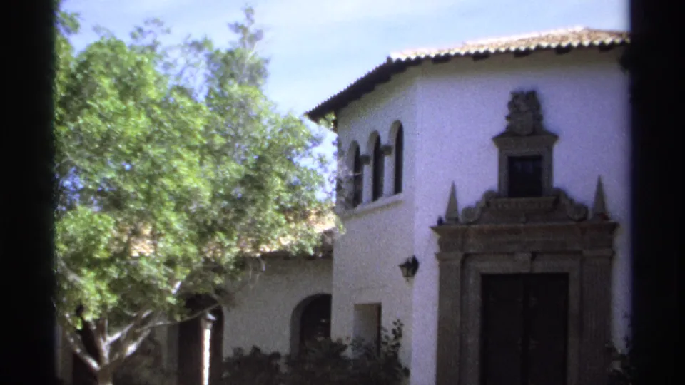 1975 Guaymas home-movie view of a white colonial-style facade partly obscured by a leafy tree and film gate edge.