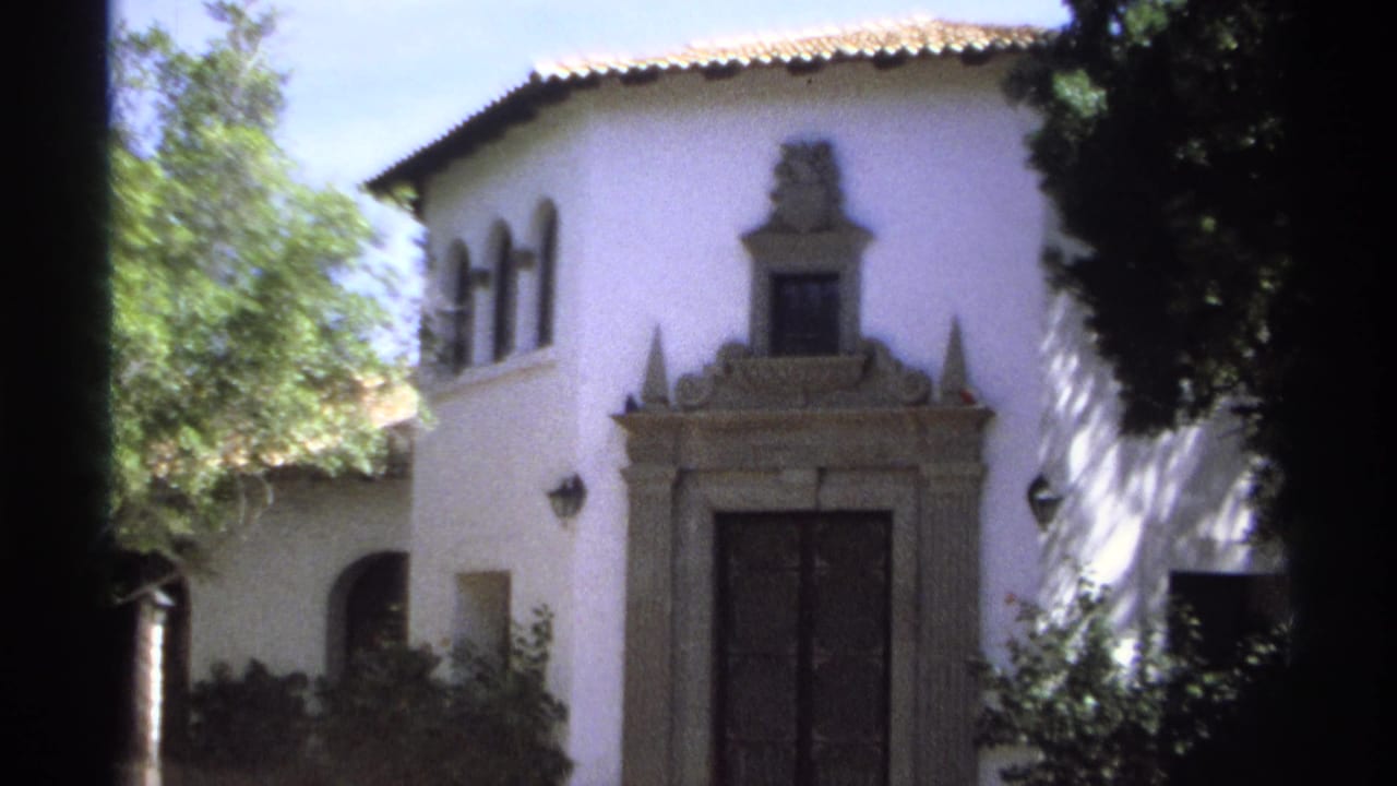 1975 Guaymas, Mexico 8mm frame showing a white stucco villa facade, decorative stone entrance, dark double doors, arched windows, shrubs, and tree shade.