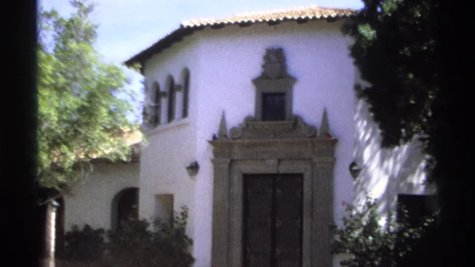 1975 Guaymas, Mexico 8mm frame showing a white stucco villa facade, decorative stone entrance, dark double doors, arched windows, shrubs, and tree shade.