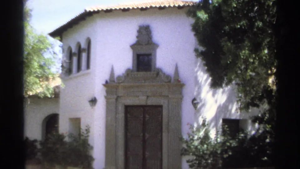 1975 Guaymas, Mexico 8mm frame of a sunlit white stucco villa with arched upper windows, an ornate stone doorway, dark doors, shrubs, and overhanging tree branches.