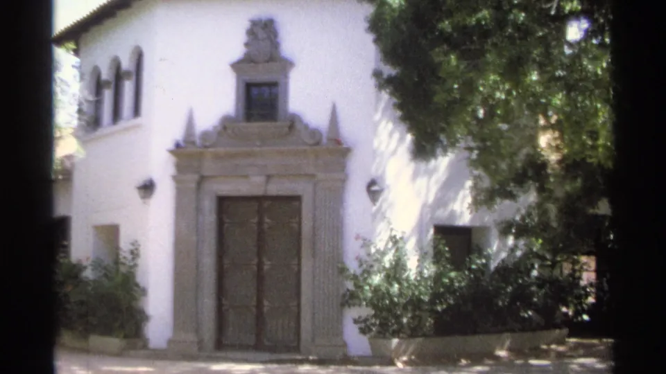 1975 Guaymas, Mexico 8mm frame showing the ornate entrance of a white stucco villa, with dark doors, arched windows, bushes, planters, and leafy shade crossing the facade.