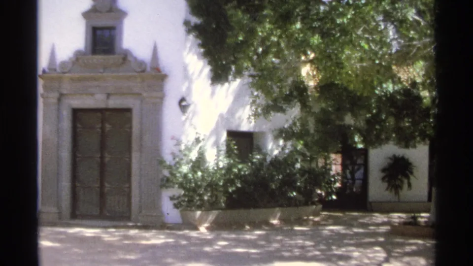 1975 Guaymas, Mexico 8mm frame of the villa front from a wider angle, showing the central decorative doorway, shaded courtyard, shrubs, trees, and a secondary door or window at right.