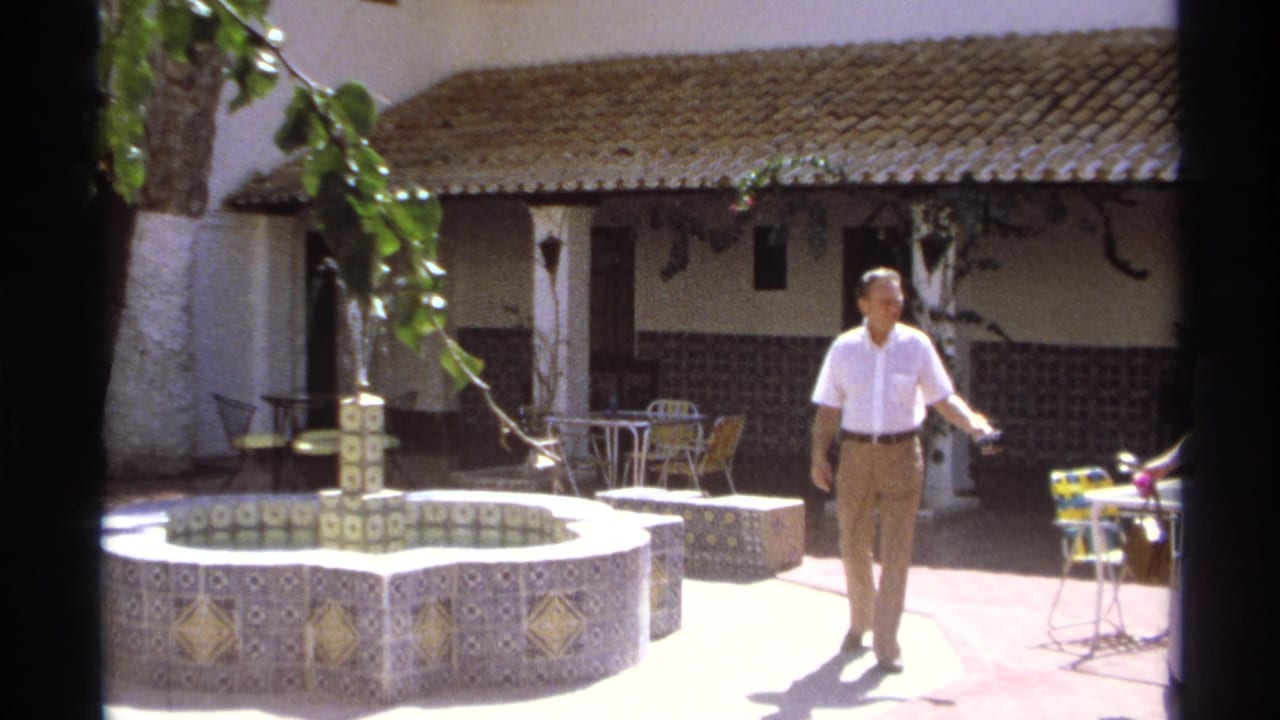 A man walks through a Guaymas courtyard near a tiled fountain, white buildings, and outdoor tables in 1975 home-movie footage.
