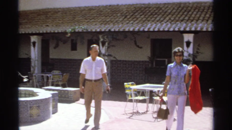 A couple walks toward the camera in a Guaymas courtyard, with patio chairs, tiled walls, and a fountain nearby.