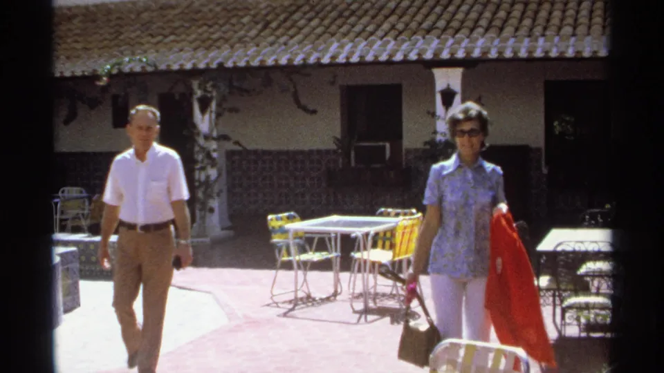 The couple continues across the Guaymas courtyard, passing yellow chairs, tables, tiled walls, and shaded doorways.
