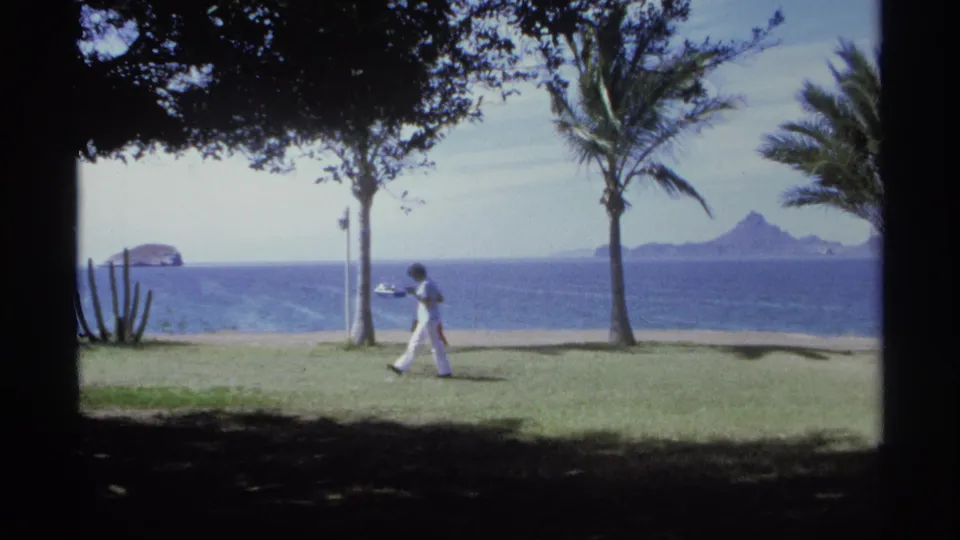 The final 1975 Guaymas frame holds on a shaded coastal park as a person walks past palms and cactus with ocean and islands beyond.