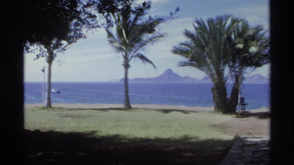 1975 8mm view of a Guaymas beach lawn with palms, a path, blue water, and distant mountains or islands.
