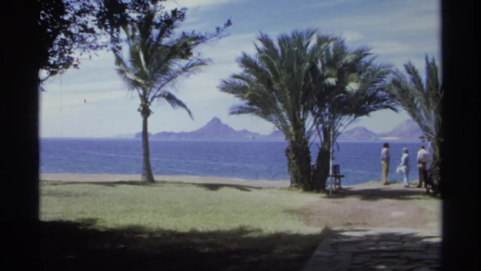 Guaymas beachside scene from 1975 with visitors near palms, a lawn, ocean water, and distant mountain shapes.