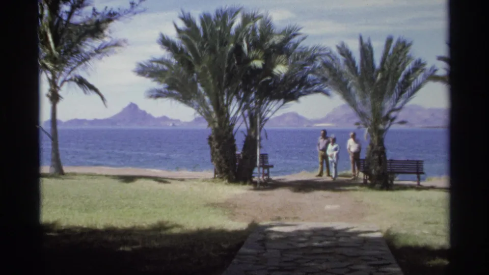 1975 Guaymas home-movie frame of three adults standing by palms and a bench along the bay, with distant mountains across the water.