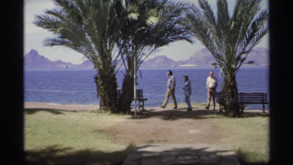1975 Guaymas frame showing three adults walking past palm trees on a seaside promenade, with benches, blue water, and mountains beyond.