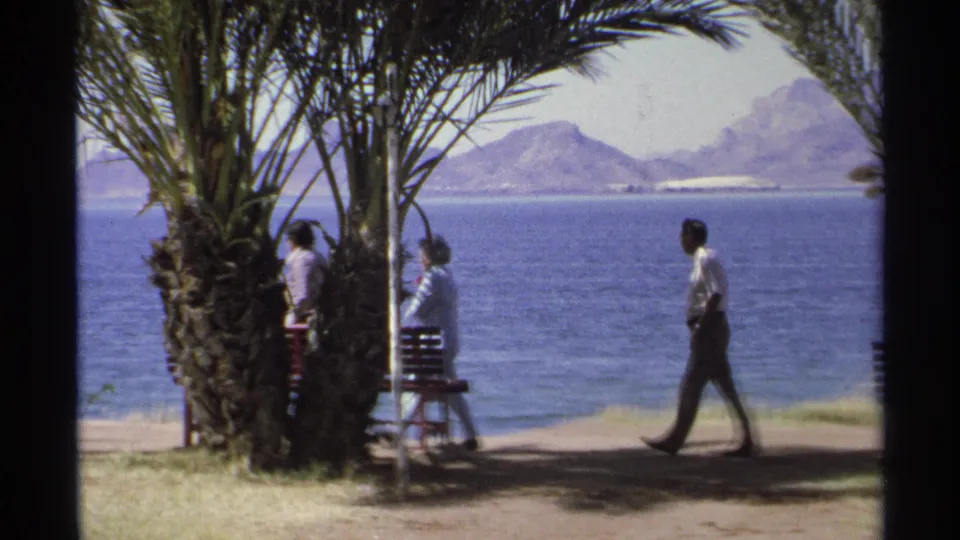Adults walk along a Guaymas waterfront promenade framed by palms, benches, blue sea, and hazy mountains in 1975 8mm footage.