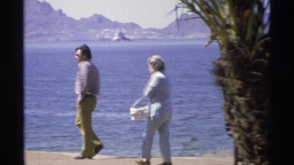 A man and woman in 1970s clothing walk along the Guaymas waterfront, with blue water, palms, and mountains in the background.
