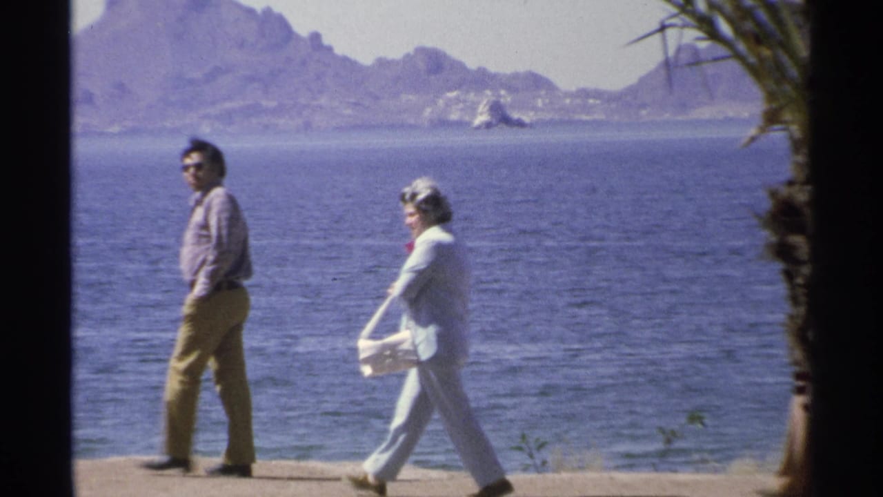 Two adults walk along a seaside path with ocean and mountains behind them.