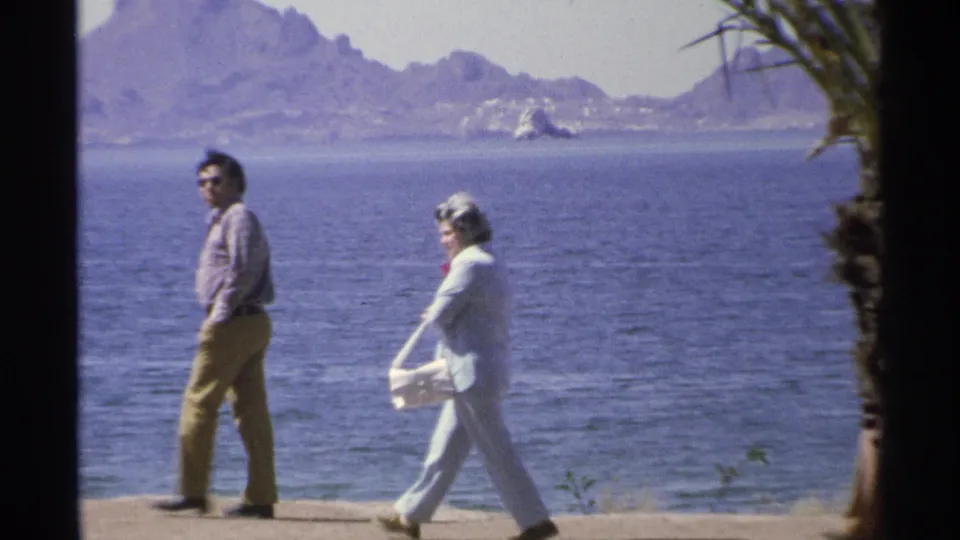 In 1975 Guaymas, Mexico, a man and woman walk along a waterfront path with blue ocean and distant mountains in view.