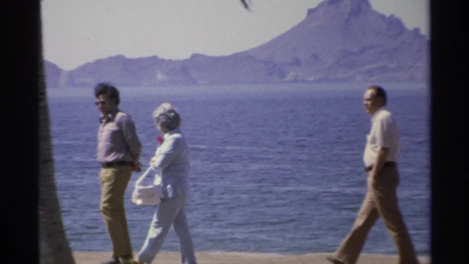 A 1975 Guaymas waterfront view shows three adults on a promenade, including a woman carrying a white purse, with ocean behind.