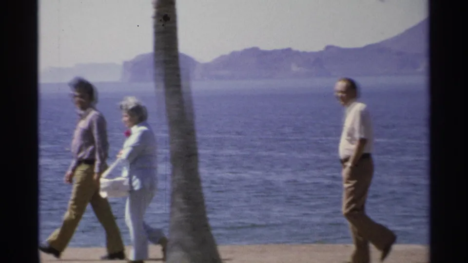 In Guaymas, Mexico, three adults appear along a coastal promenade, with a palm trunk, blue water, and mountains visible.