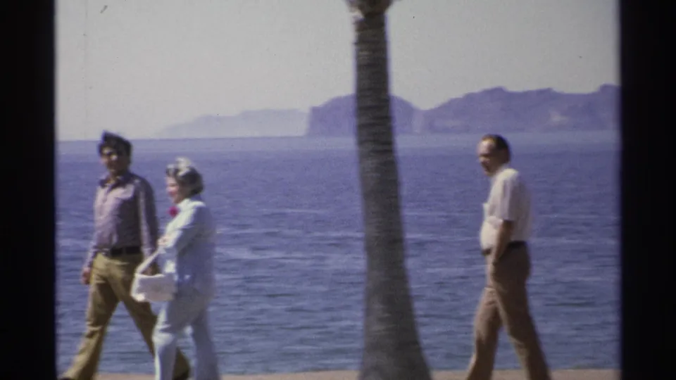 In 1975 Guaymas, Mexico, three adults walk along a seaside promenade with ocean, palm trunk, and hazy hills in the background.