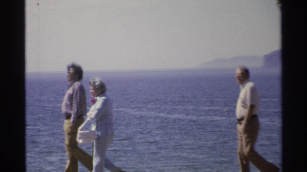 Adults walking along a waterfront path with blue ocean and mountains in the distance.