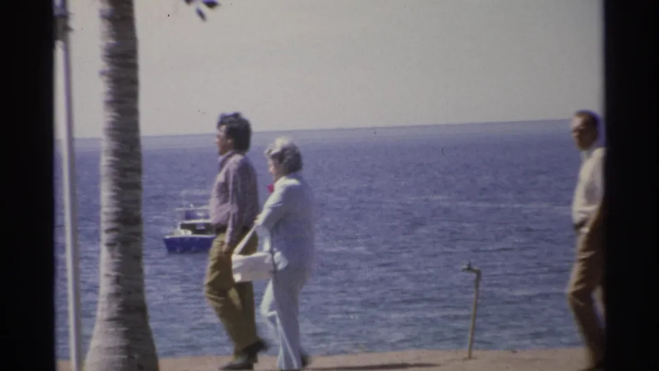 On the Guaymas seaside promenade, three adults walk past a palm trunk while a small boat appears offshore.