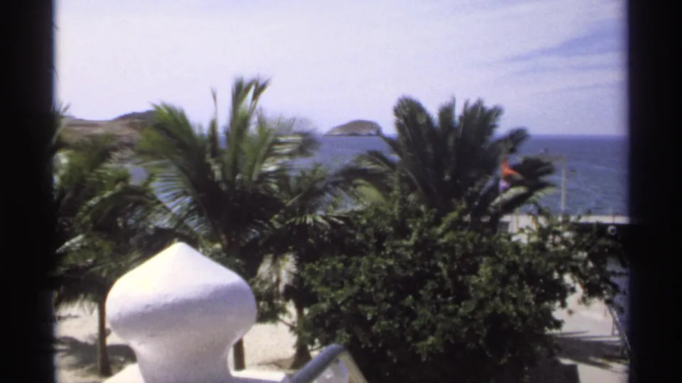 1975 Guaymas beach view with palms, a white domed foreground feature, ocean, a dock area, and tiny distant figures near the shoreline.