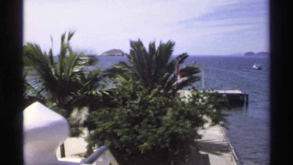 Guaymas, Mexico 8mm frame showing palms, shoreline, a dock with a flag, open ocean, distant islands, and a small boat offshore.
