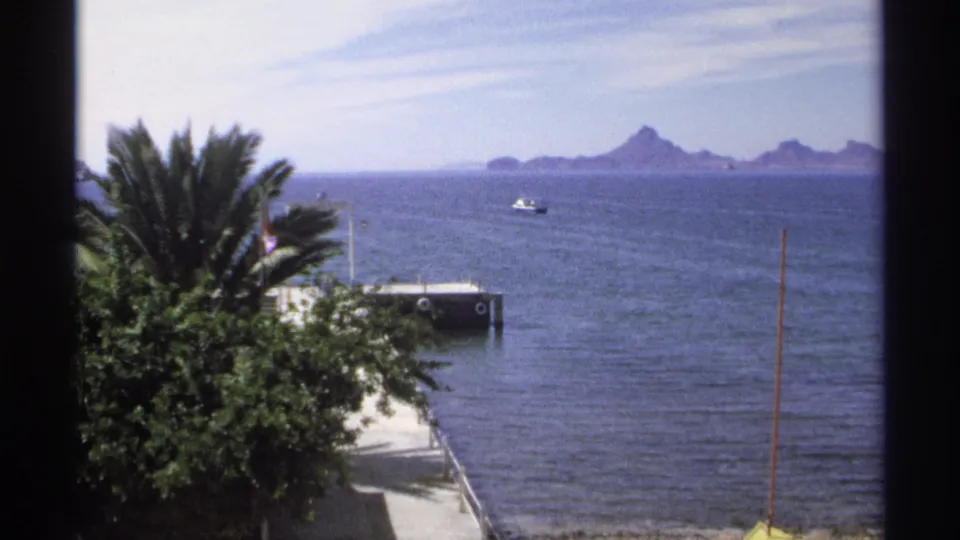 Guaymas seaside home-movie frame showing palms and shrubs near a pier, with a small boat and jagged islands under a bright sky.