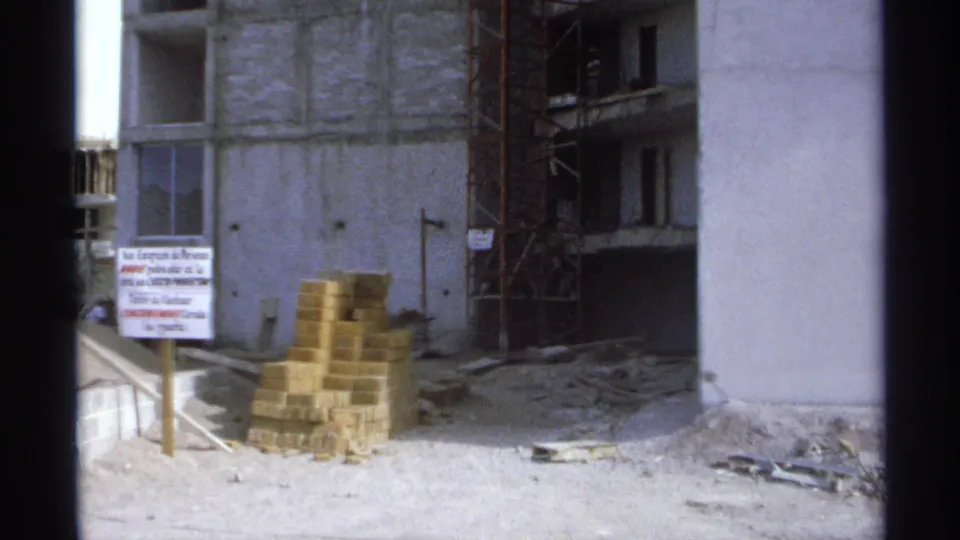 Fixed 8mm view of a Guaymas construction site showing concrete columns, scaffolding, brick stacks, and a Spanish sign.