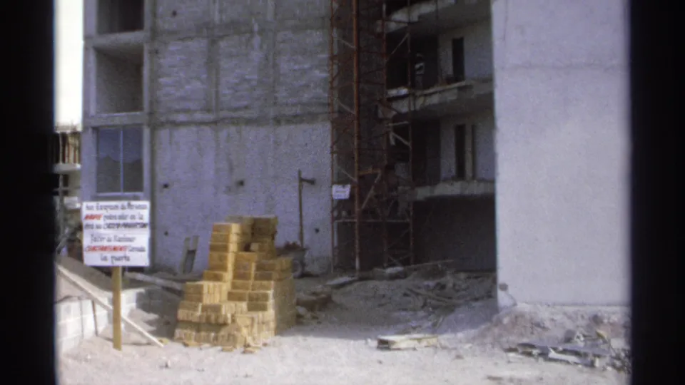 1975 home-movie frame of the Guaymas building site, with stacked bricks, scaffolding, rubble, and a Spanish sign near the entrance.