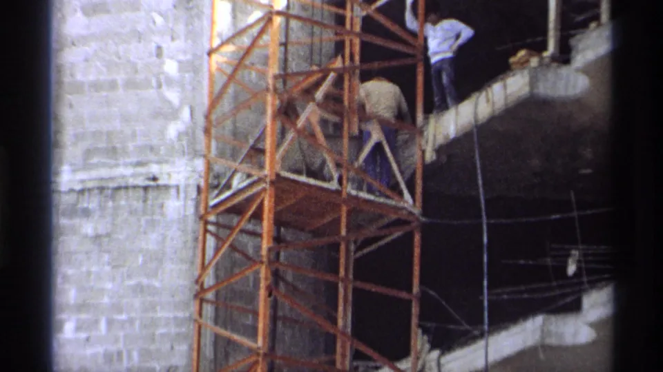 Workers appear on orange scaffolding at a 1975 Guaymas construction site beside an unfinished concrete wall and open building edge.