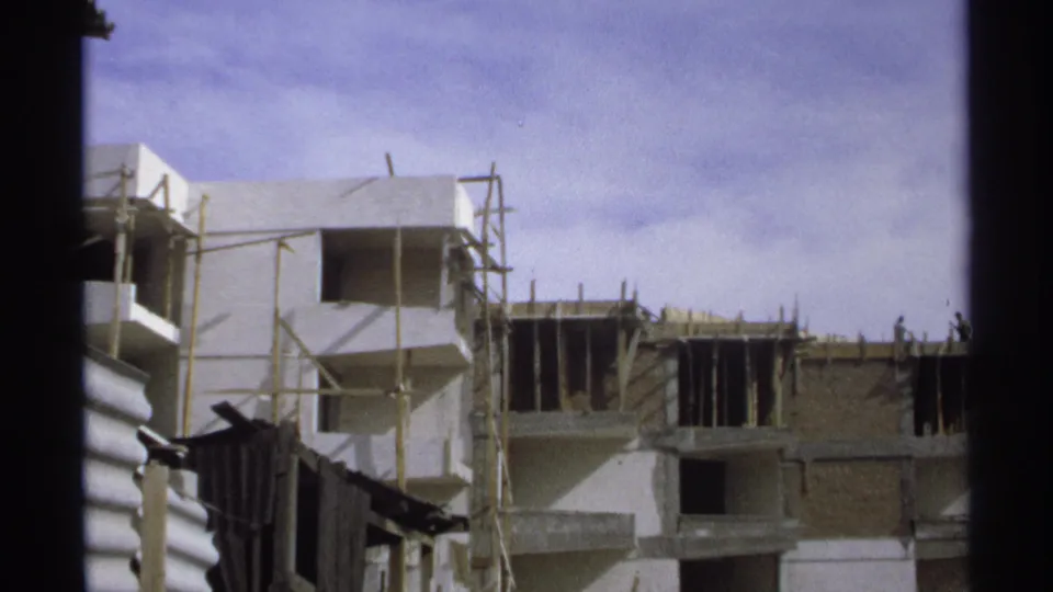 Wider 1975 Guaymas view showing unfinished concrete buildings, scaffold poles, open window bays, and a bright partly cloudy sky.