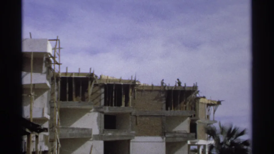 1975 Guaymas, Mexico 8mm view of a cropped construction site, workers on the top level, and palm fronds in the foreground.