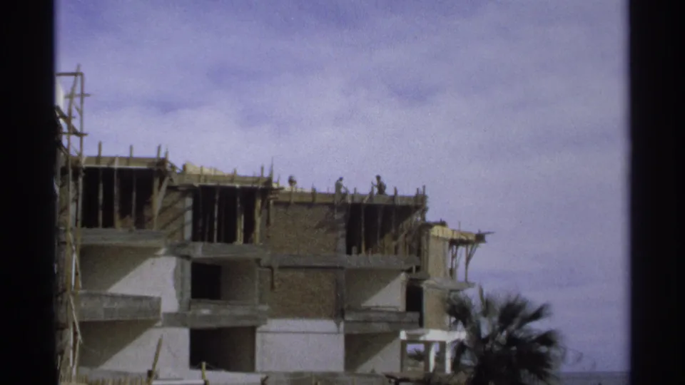 1975 Guaymas, Mexico frame showing workers on an unfinished multi-story building, with a palm tree at lower right.