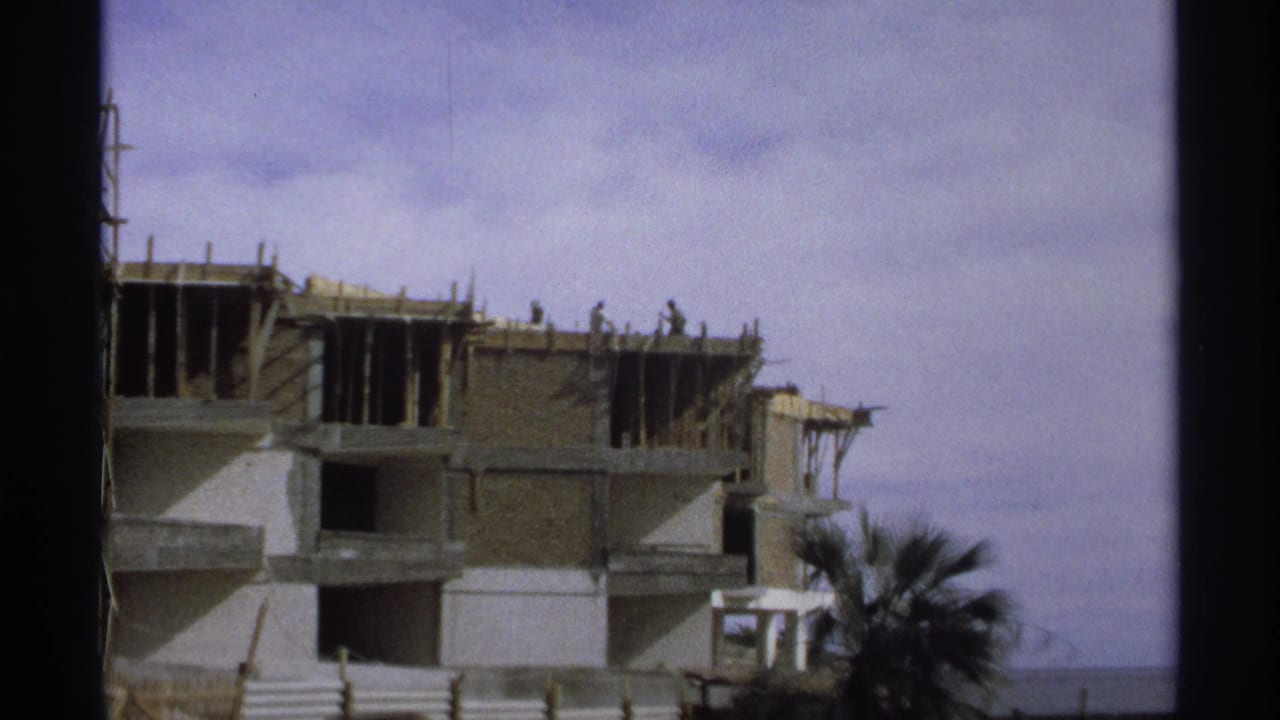 1975 Guaymas, Mexico 8mm frame showing a multi-story building under construction with workers, scaffolding, palm fronds, and ocean beyond.